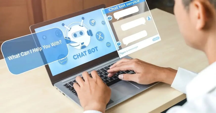 A person typing on a laptop with a chatbot interface displayed on the screen. The chatbot has a robotic icon and text bubbles, including a message saying, 'What can I help you with?'