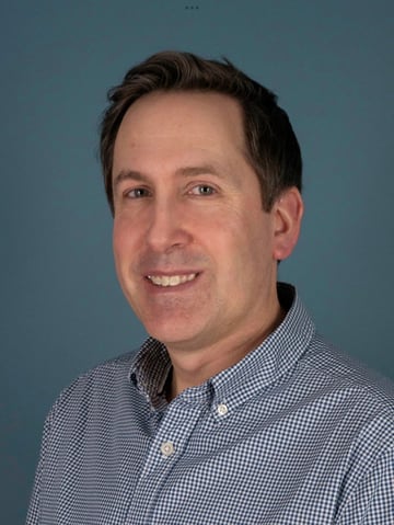 A professional headshot of a smiling man with short brown hair, wearing a blue and white checkered button-up shirt, against a solid blue background. He appears to be looking directly at the camera in a well-lit portrait setting